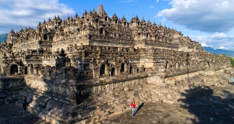 Ground-level perspective of Borobudur's intricate stone stupas with a visitor in red standing in the vast courtyard.