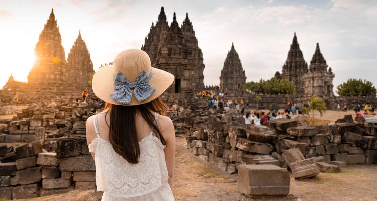 Woman in a sun hat admires Prambanan Temple at sunset, warm light illuminating ancient stone ruins.