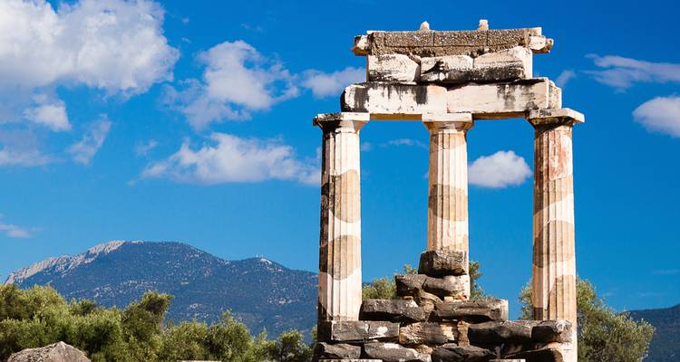 Ruina antigua de tres columnas del Tholos de Delfos con un fondo de montaña y cielo azul.