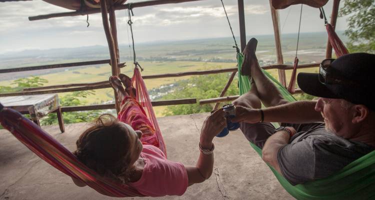 Un homme et une femme se détendent dans des hamacs colorés suspendus à une structure en bois qui surplombe une vaste vallée rurale sous un ciel dégagé.