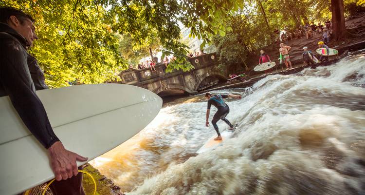 Les surfeurs chevaucent la vague stationnaire de la rivière sous des arbres feuillu tandis que les spectateurs observent depuis un pont