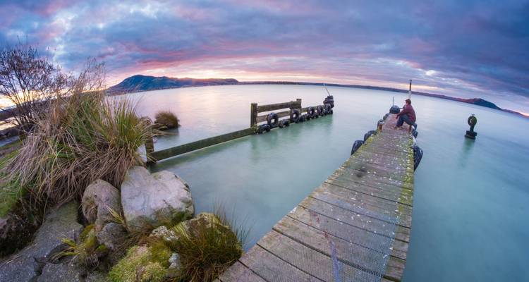 Personne assise au bout d'une jetée en bois au-dessus d'un lac calme pendant un lever de soleil coloré avec des nuages doux et des collines lointaines.