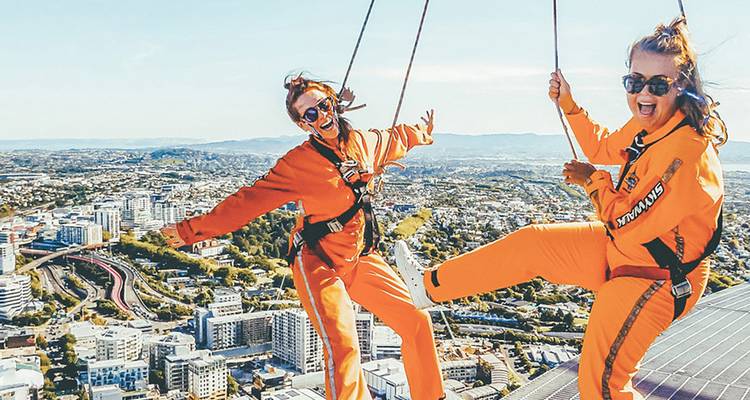 Aventureros en monos naranjas se equilibran en una pasarela aérea de gran altitud con vista a una ciudad y puerto.