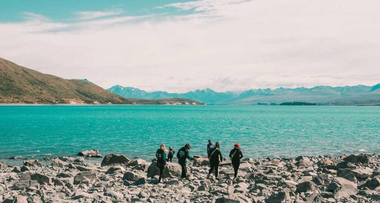 Un pequeño grupo se encuentra en la orilla rocosa de un lago alpino de color turquesa con montañas nevadas en la distancia.