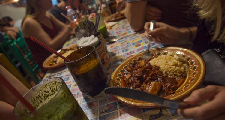 Una mesa animada en un restaurante con comensales disfrutando de un plato de carne guisada con arroz, rodeados de bebidas y decoración colorida.