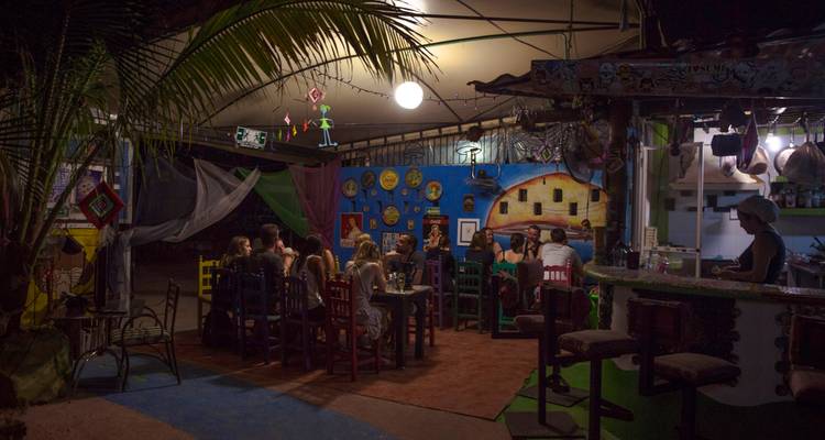 Un comedor al aire libre y con poca luz donde un grupo de viajeros se sienta en mesas de madera coloridas bajo un techo de hojas de palma.