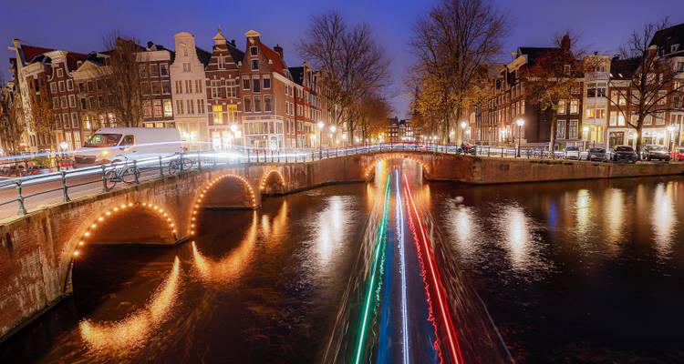 Nachtansicht eines Amsterdamer Kanals mit Lichtspuren von Booten und beleuchteten Brücken, die sich im Wasser spiegeln