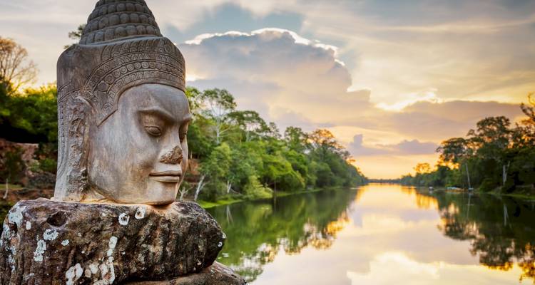 Close-up of a stone Buddha statue overlooking a river at sunset.