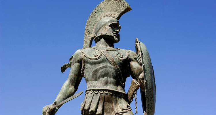 Statue of a warrior with a spear and shield against a clear blue sky.