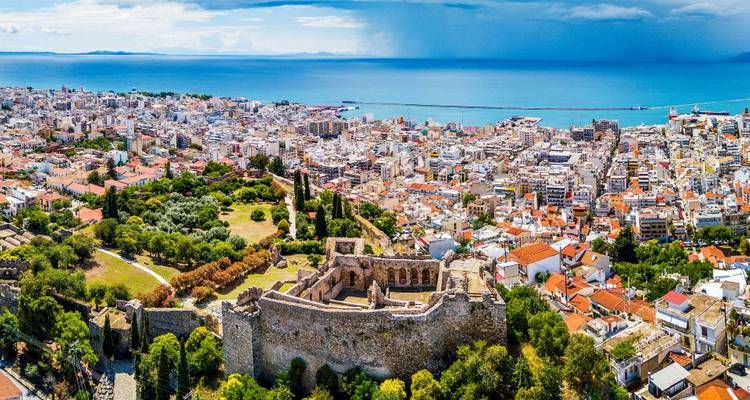 Ancient ruins of a city with buildings and the sea in the background.