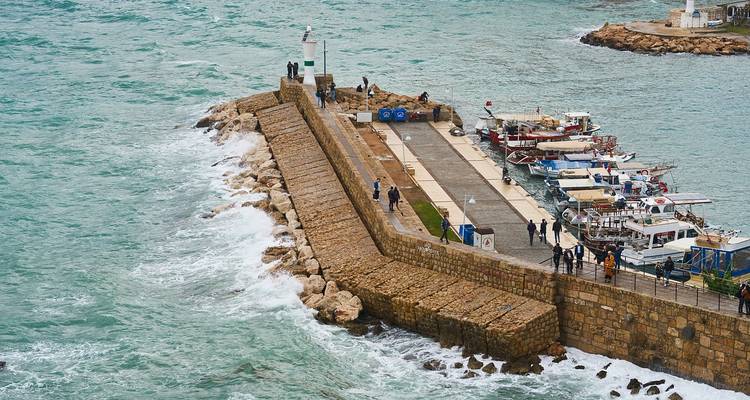 Pier with people and moored boats as waves crash against the rocks.