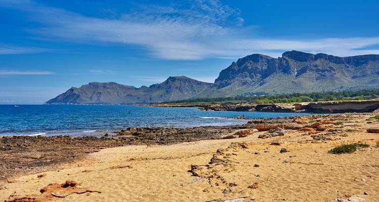Plage rocheuse avec des montagnes au loin sous un ciel bleu.