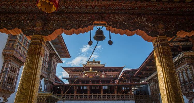 Arco de madera decorativo y campana colgante que enmarcan una vista hacia un patio de dzong butanés.