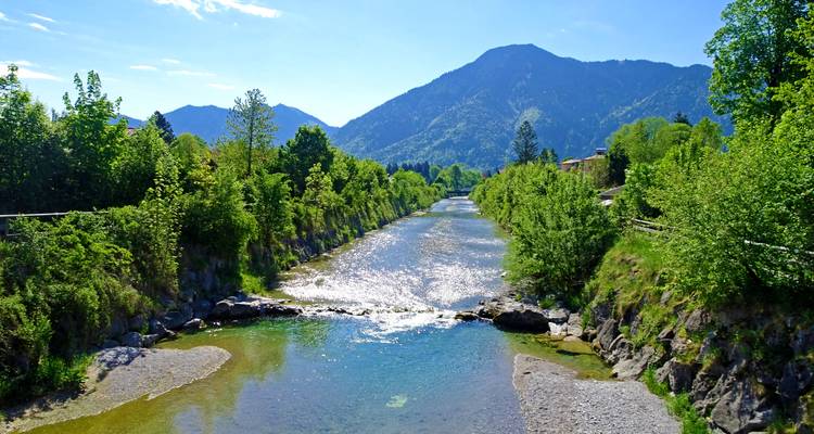 Rivière de montagne cristalline s'écoulant entre des rives verdoyantes avec des pics boisés en arrière-plan par une journée ensoleillée.