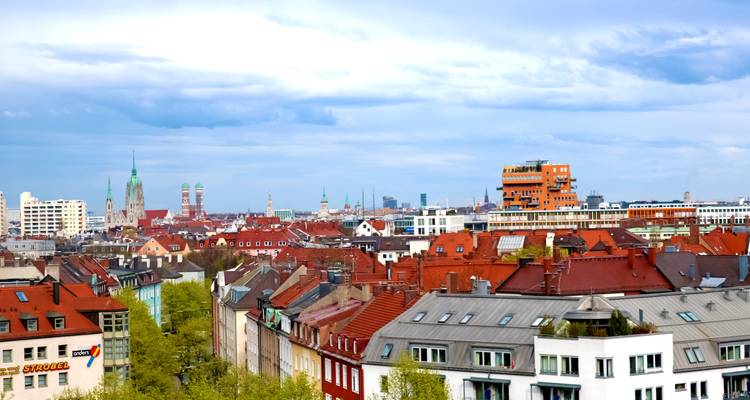 Panoramablick auf die Münchner Skyline mit roten Dächern, Kirchtürmen und modernen Gebäudekomplexen unter dramatischen Wolken.
