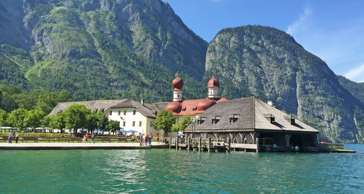 Picturesque lakeside chapel with red onion domes set against towering green alpine cliffs and emerald water.