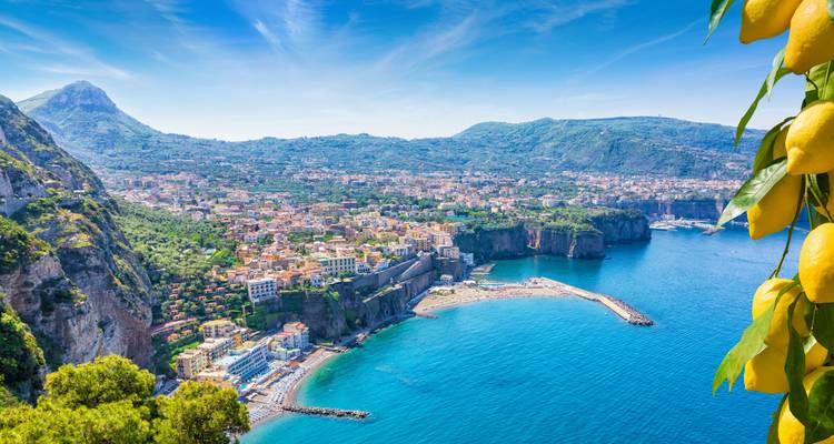 Vista costera de Sorrento con acantilados, edificios y un mar azul. Limones cuelgan en primer plano.