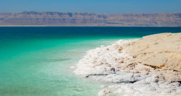 Leuchtend türkisfarbenes Wasser einer salzkrustenbedeckten Küstenlinie, das auf ein tiefblaues Meer trifft, mit Wüstenbergen in der Ferne.