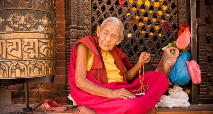 Elderly monk in crimson robe sits praying with mala beads beside ornate prayer wheel.