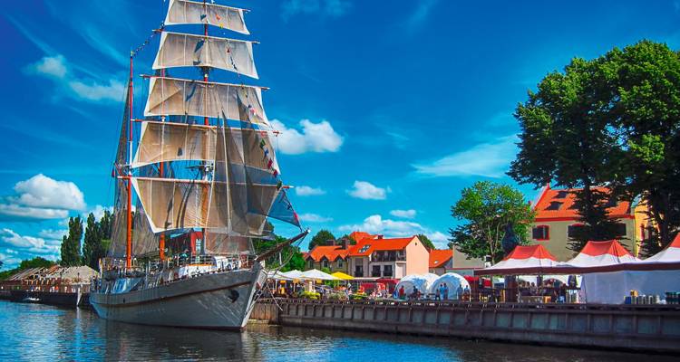 Ein großes Segelschiff, das in der Nähe einer Uferpromenade mit Zelten und Gebäuden angelegt hat.