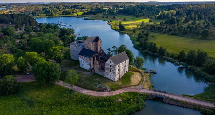 Ein mittelalterliches Steinschloss befindet sich auf einer kleine bewaldeten Insel inmitten eines ruhigen Sees und einer sanfthügeligen grünen Landschaft.