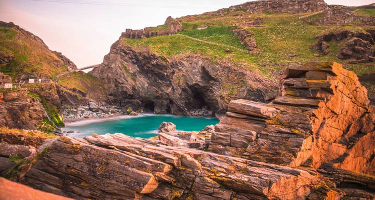 Rocky cliffs surround a small turquoise cove with a stone bridge visible in the distance at dusk.