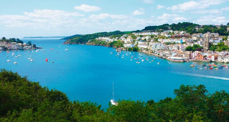 A picturesque harbor town with whitewashed houses stretches along a vivid blue estuary dotted with colorful sailboats.