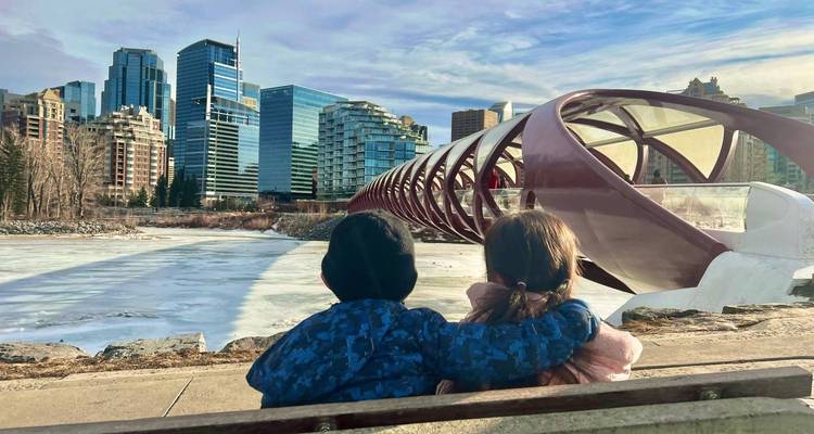 Two children sitting by a river with modern city architecture in the background.