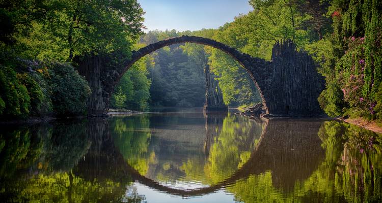 Un pont en arche parfait se reflétant dans une eau calme entourée de forêt.
