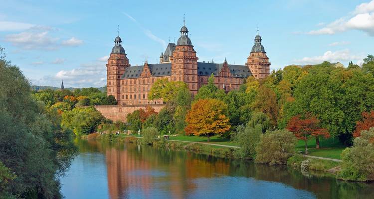 Majestic castle rises above autumn-colored trees beside a tranquil river under blue skies.