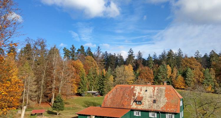 Rustic house with red-tiled roof set against colourful autumn forest under blue sky.