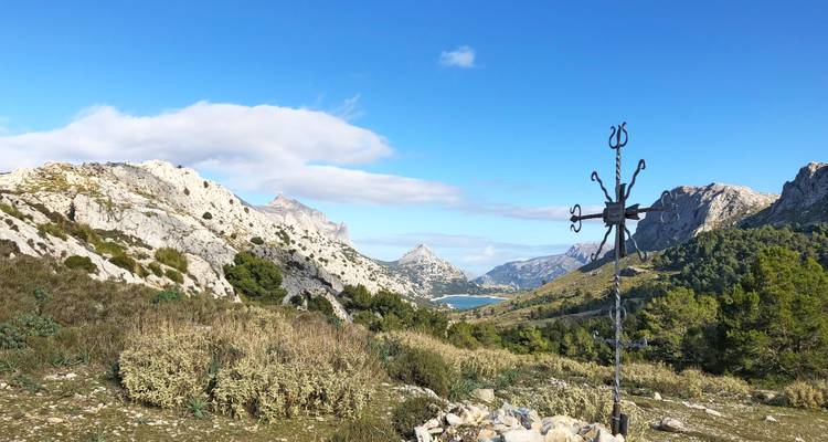 Panoramisches Bergtal mit schroffen Kalksteingipfeln, Stausee und eiserner Gipfelkreuz unter blauem Himmel