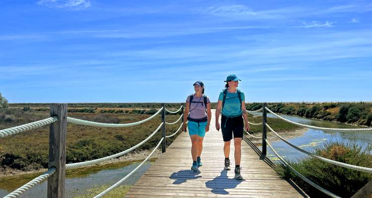 Zwei Wanderer überqueren einen hölzernen Steg über Feuchtgebiete unter dem strahlend blauen Himmel der Algarve