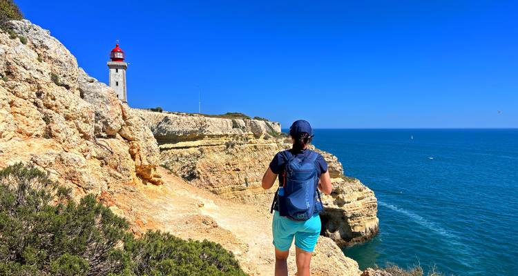 Ein einsamer Wanderer steht auf goldenen Meeresklippen und blickt auf einen Leuchtturm mit roter Spitze und den tiefblauen Ozean.