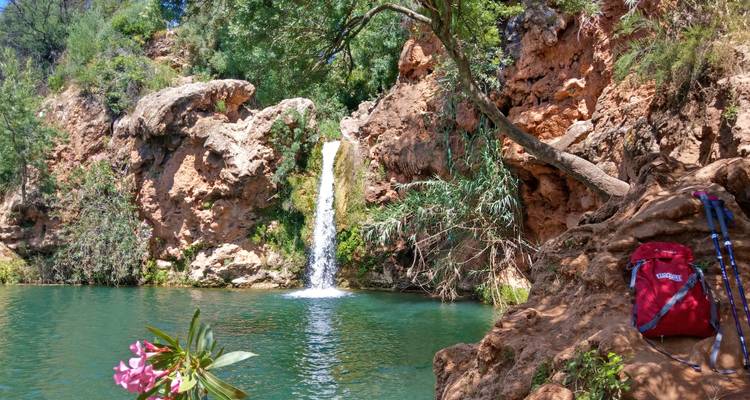 Ein kleiner Wasserfall fällt in einen klaren Tümpel, der von roten Felsklippen und Grün umgeben ist, mit Wanderausrüstung, die in der Nähe liegt.