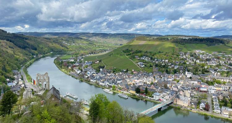 Zwevend luchtbeeld van een Moezelstadje met kasteelruïnes, bruggen en boten langs de kronkelende rivier.