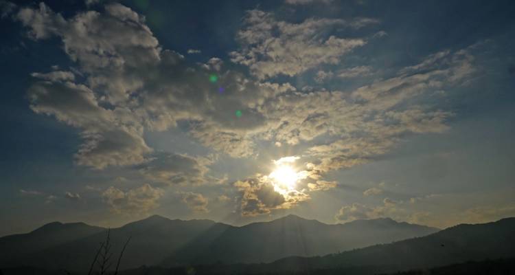 Zon die door wolken heen gluurt boven bergen.