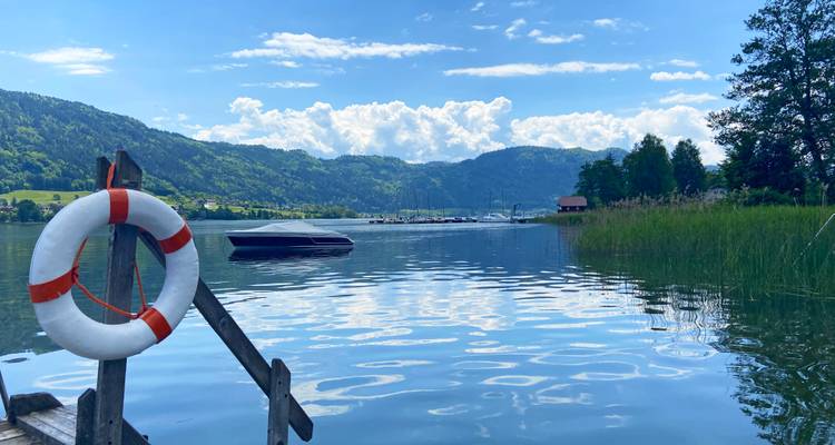 Ein ruhiger See, umgeben von bewaldeten Hügeln, mit einem Rettungsring, der an einem Holzsteg im Vordergrund hängt.