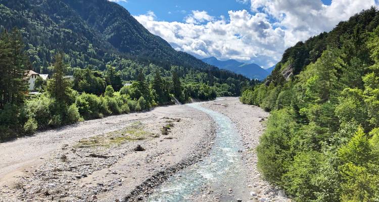 Ein steiniges alpines Flusstal schlängelt sich zwischen bewaldeten Hängen unter einem Himmel mit Kumuluswolken.