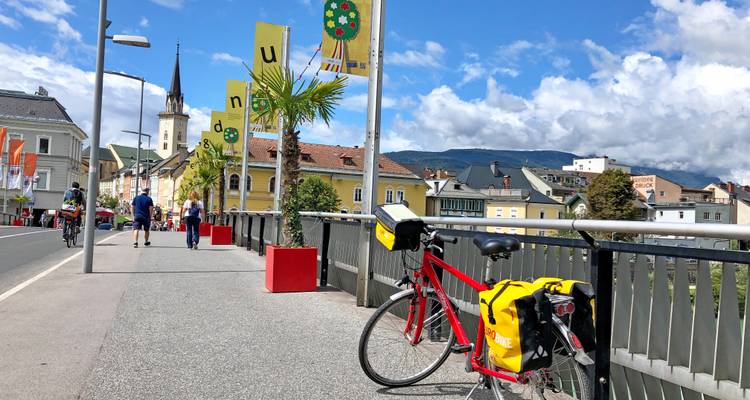 Ein rotes Tourenrad mit Packtaschen lehnt an einem Brückengeländer in einer kleinen österreichischen Stadt mit Bergen im Hintergrund.