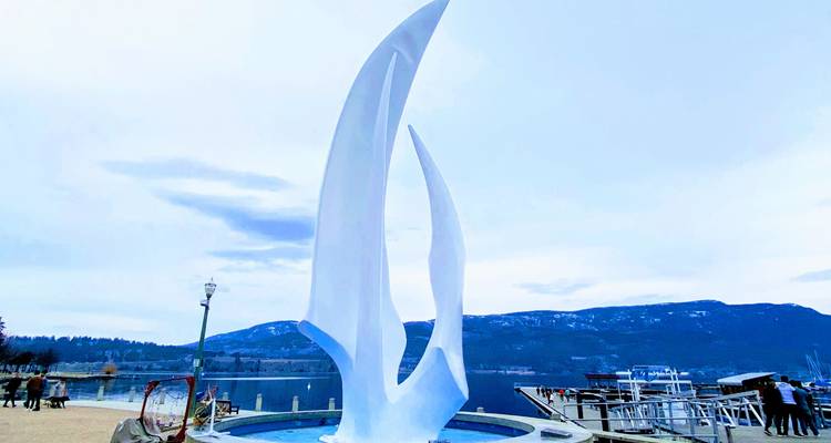 Tall white sculpture by a waterfront with mountains in the distance.