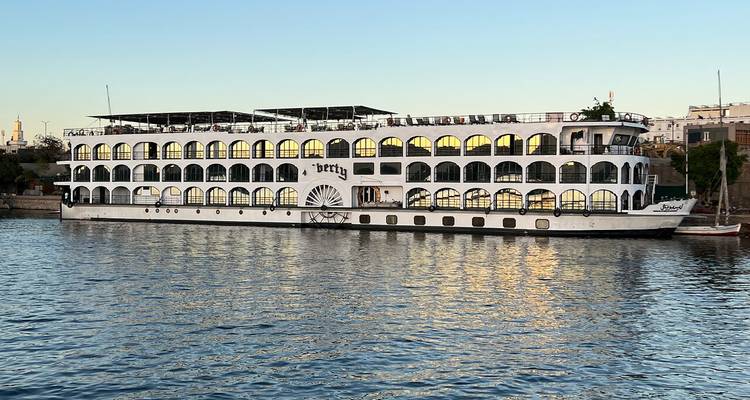 Een groot wit cruiseschip aangemeerd aan een kalme rivier, met reflecties zichtbaar in het water.