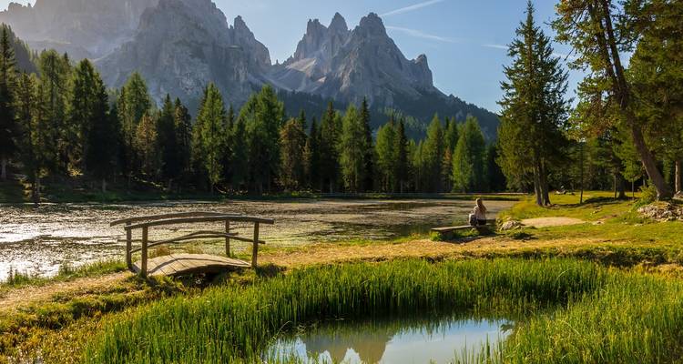 Pont en bois rustique et une personne près d'un lac tranquille avec des montagnes en arrière-plan.