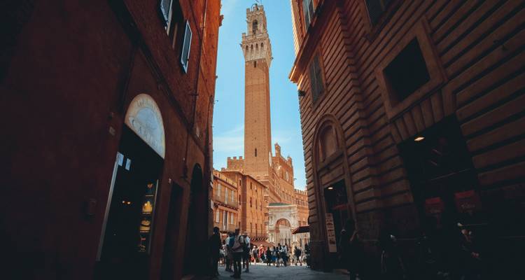Eine Straßenansicht in Siena mit gehenden Menschen, die historische Gebäude und einen hohen Turm zeigt.
