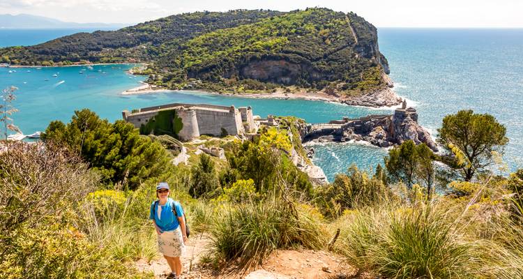 Hiker on a trail overlooking a coastal fortress and blue sea.