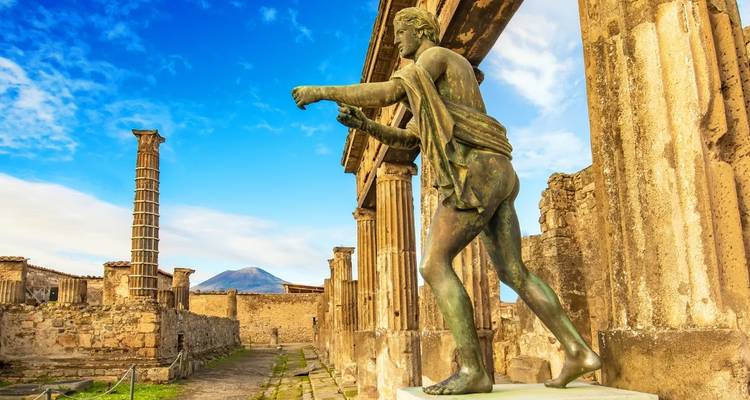 Bronzestatue in antiken Pompeji-Ruinen mit dem Vesuv im Hintergrund unter blauem Himmel.
