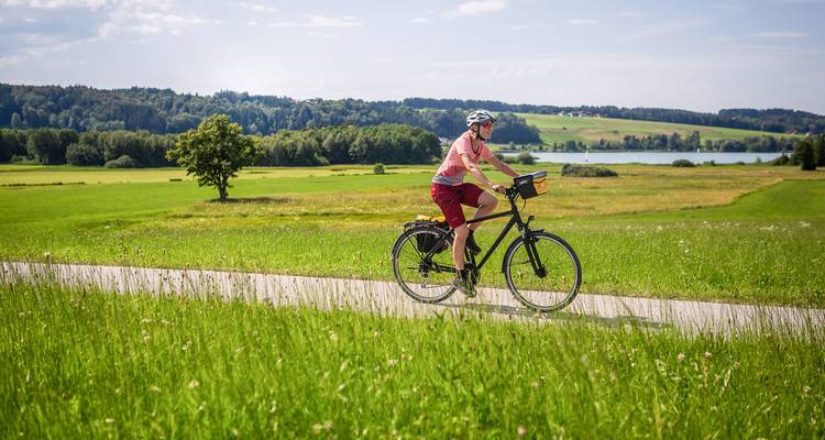 Een eenzame fietser rijdt over een glad pad door glooiende groene velden met een klein meer en beboste heuvels in de verte.