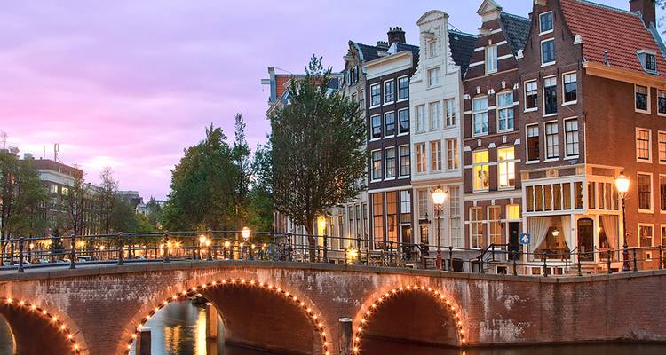 Le pont en arche du canal d'Amsterdam illuminé de guirlandes lumineuses au crépuscule, bordé de maisons classiques à pignons sous un ciel rose