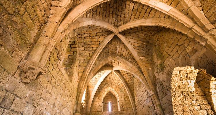 Vaulted stone arches and intersecting ribs of an ancient Crusader hall illuminated by warm light.