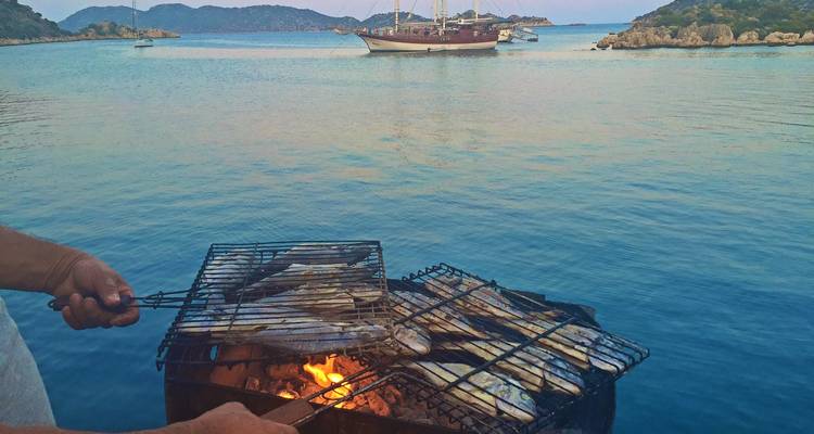 Grilling fish on a barbecue with sailboats and a serene sea in the background.